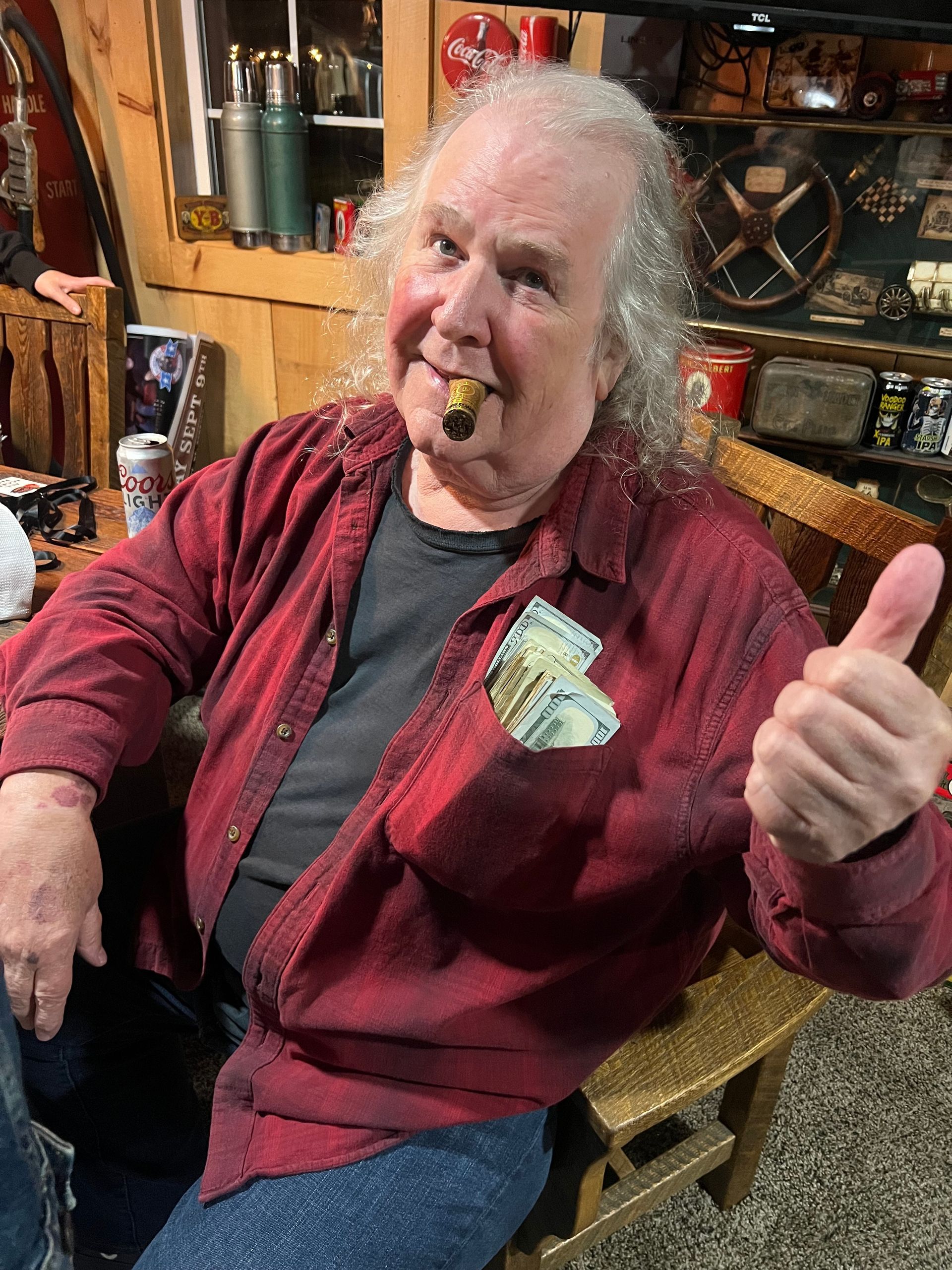 Person in a red shirt giving a thumbs-up and holding a cigar in a cozy, cluttered room.