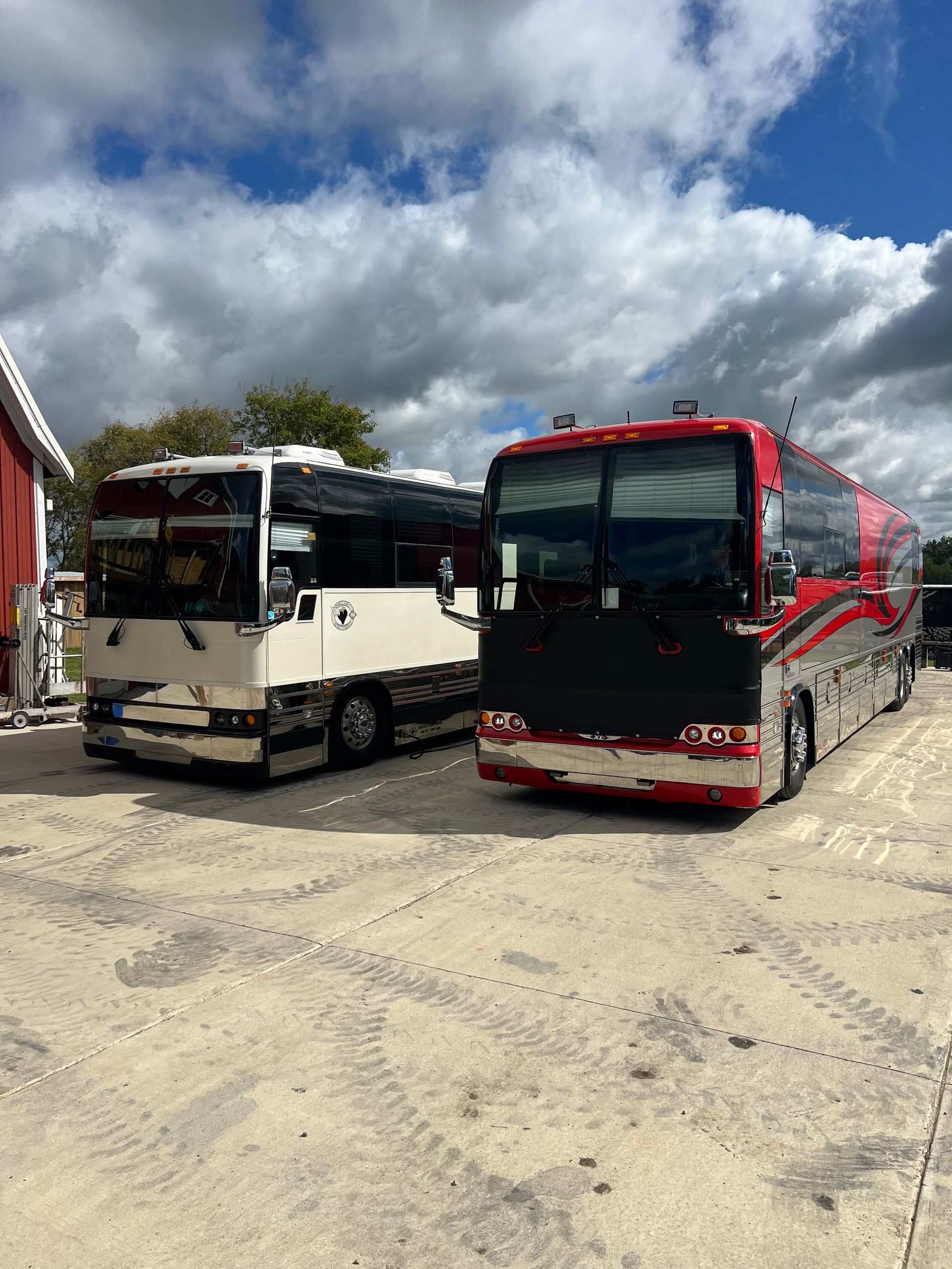 Two parked buses, one white and one red, on a gravel lot under a cloudy sky