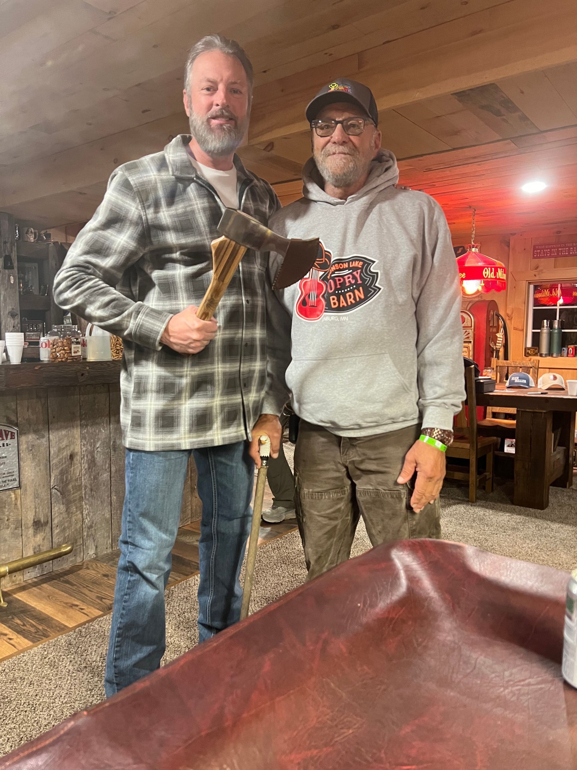Two men standing indoors, one holding a hatchet, in a rustic bar or lodge setting.