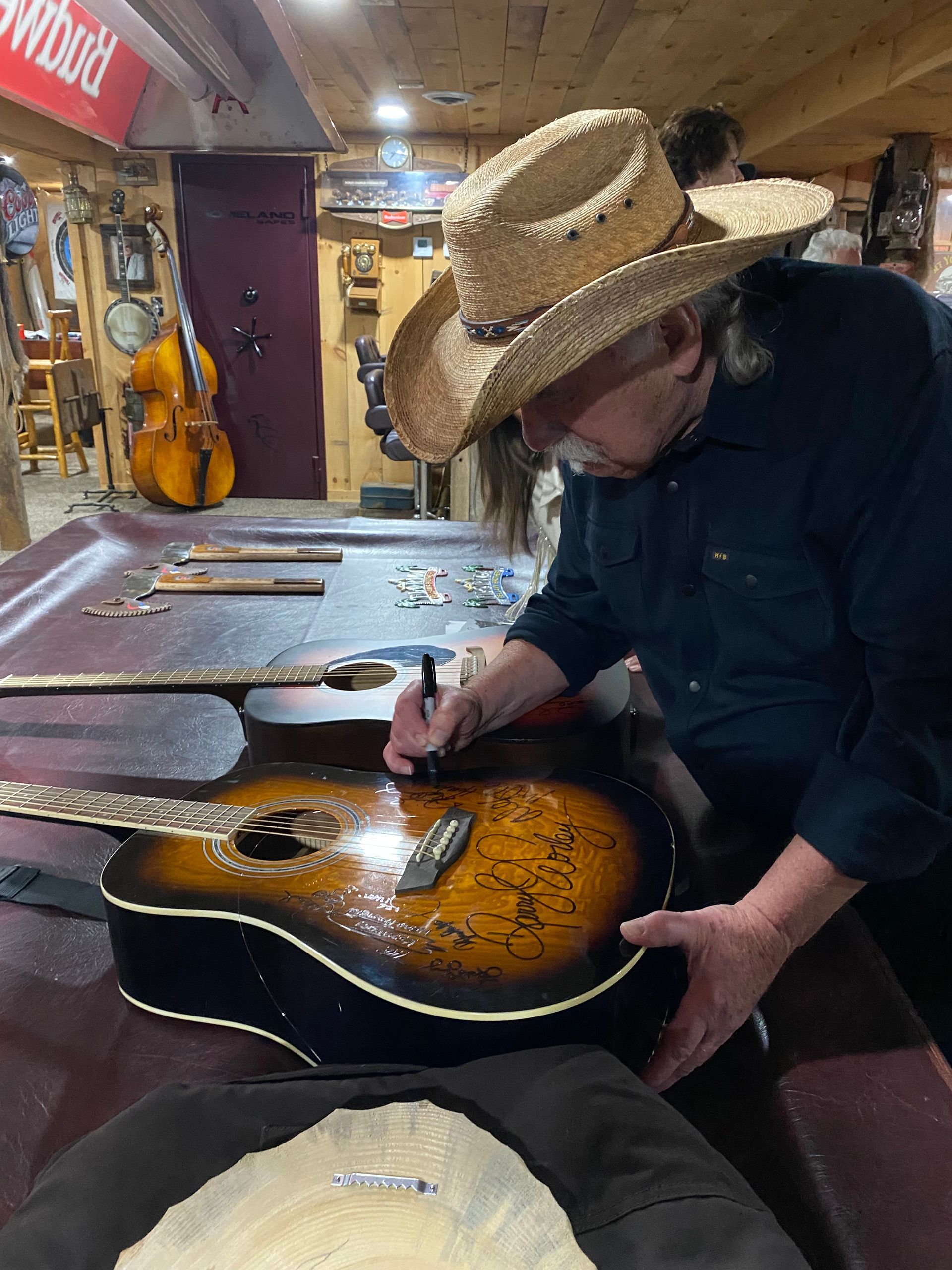 Person in a straw hat signing a sunburst acoustic guitar in a workshop, with guitars hanging on the wall.