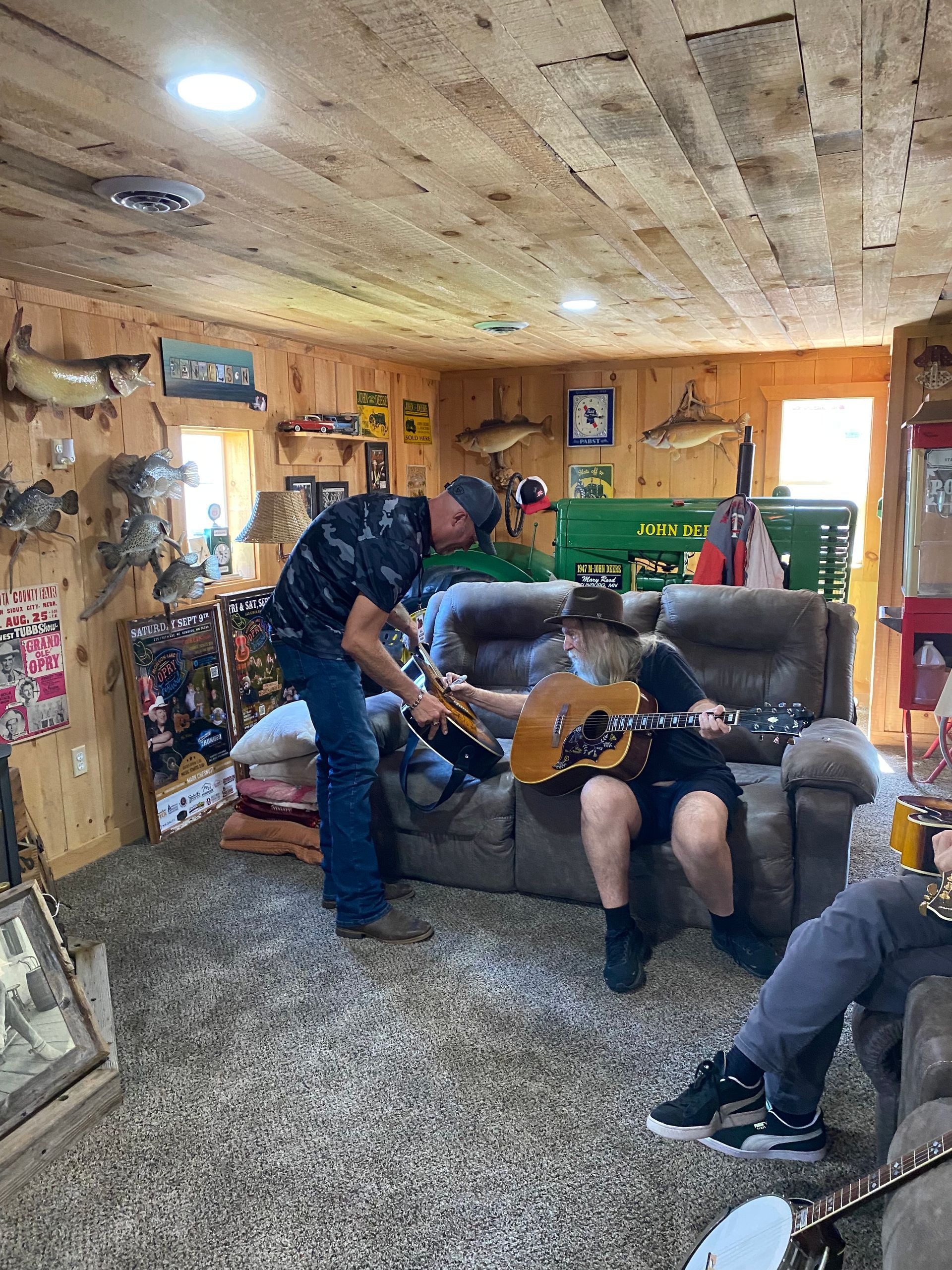 Man handing a guitar to a seated person in a cozy wood-paneled living room with a couch and pool table