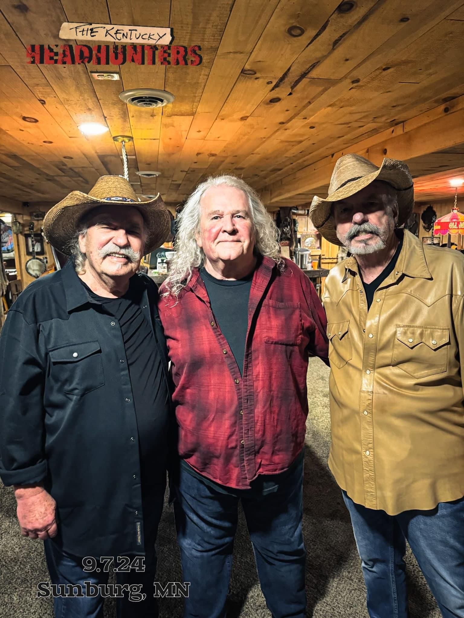 Three men in cowboy hats pose indoors under a wooden ceiling, one in a red plaid vest.