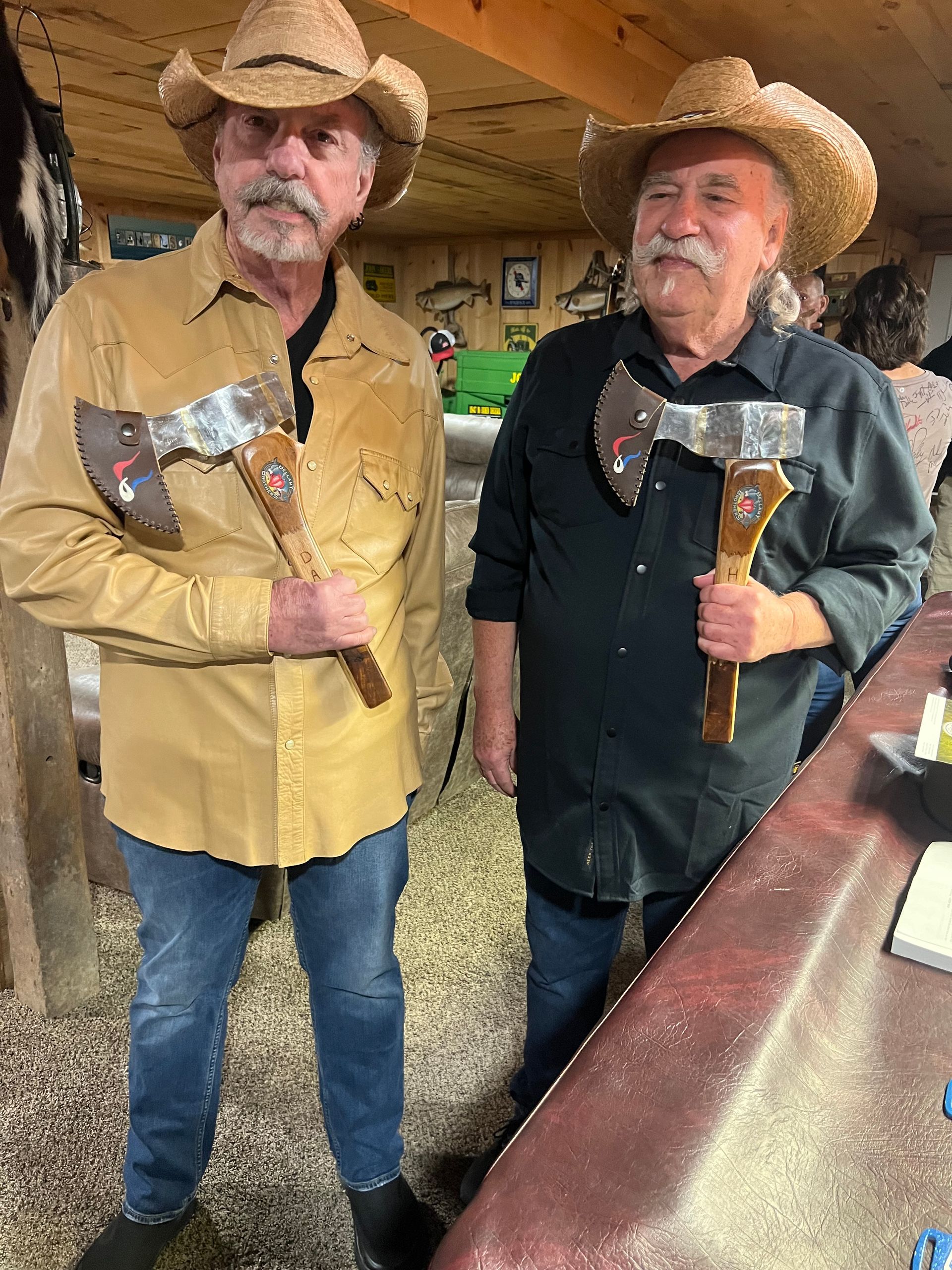 Two men in cowboy hats holding axes, standing in a rustic outdoor setting.