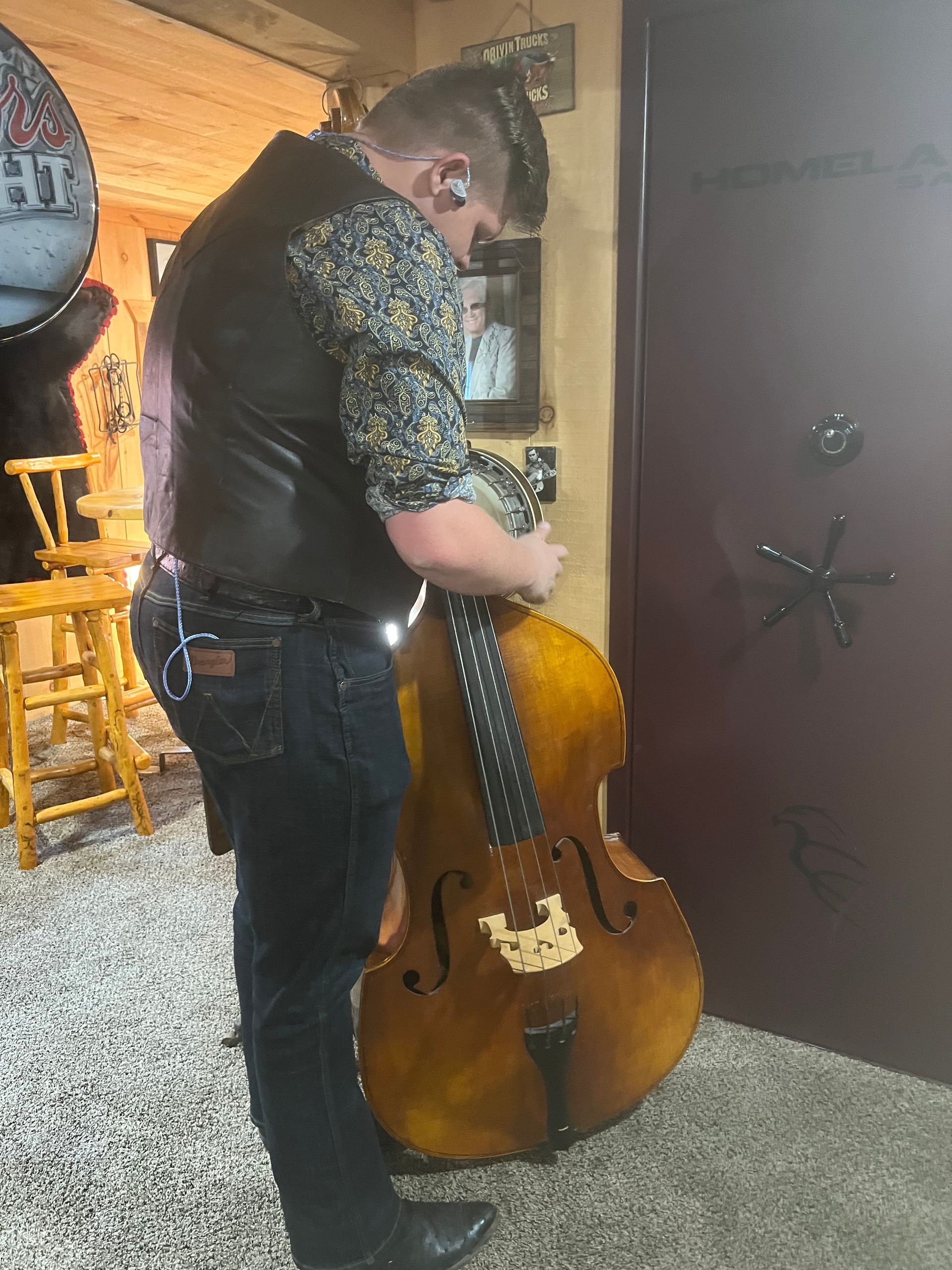 Person holding a cello in a room, standing beside a door with a carpeted floor and wooden furniture.