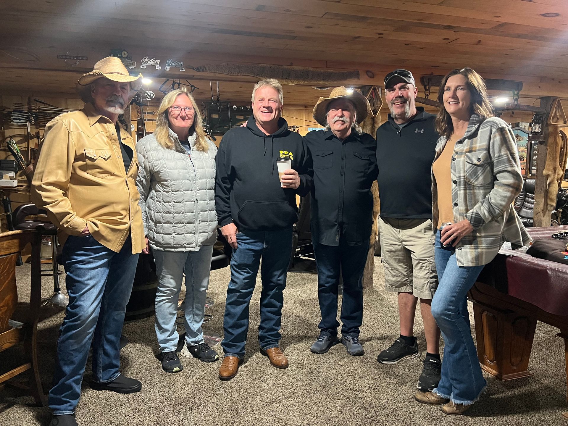 Six people standing together in a rustic wooden room, smiling at the camera with drinks in hand.