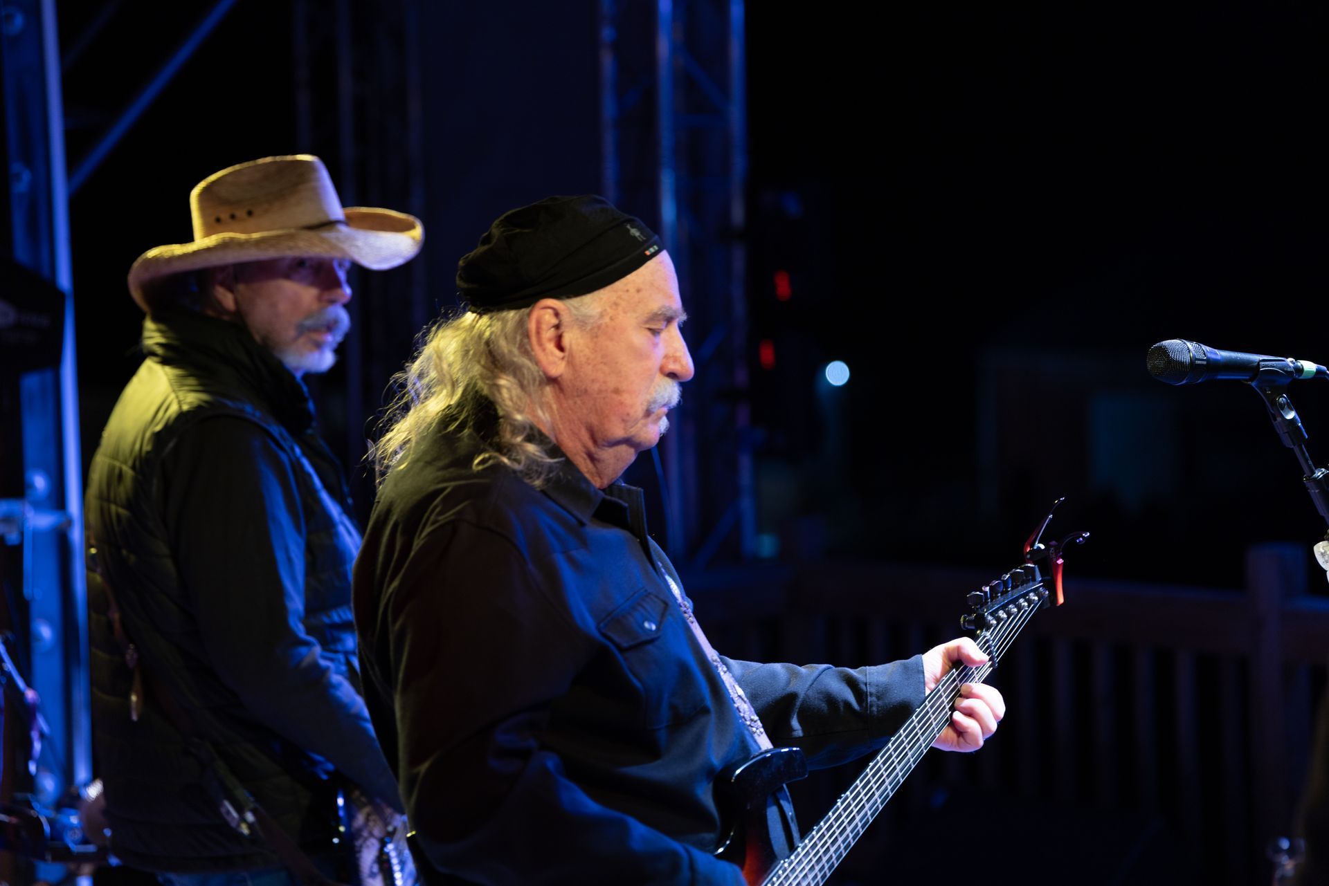 Two musicians in black outfits performing on stage, one playing guitar under blue lighting