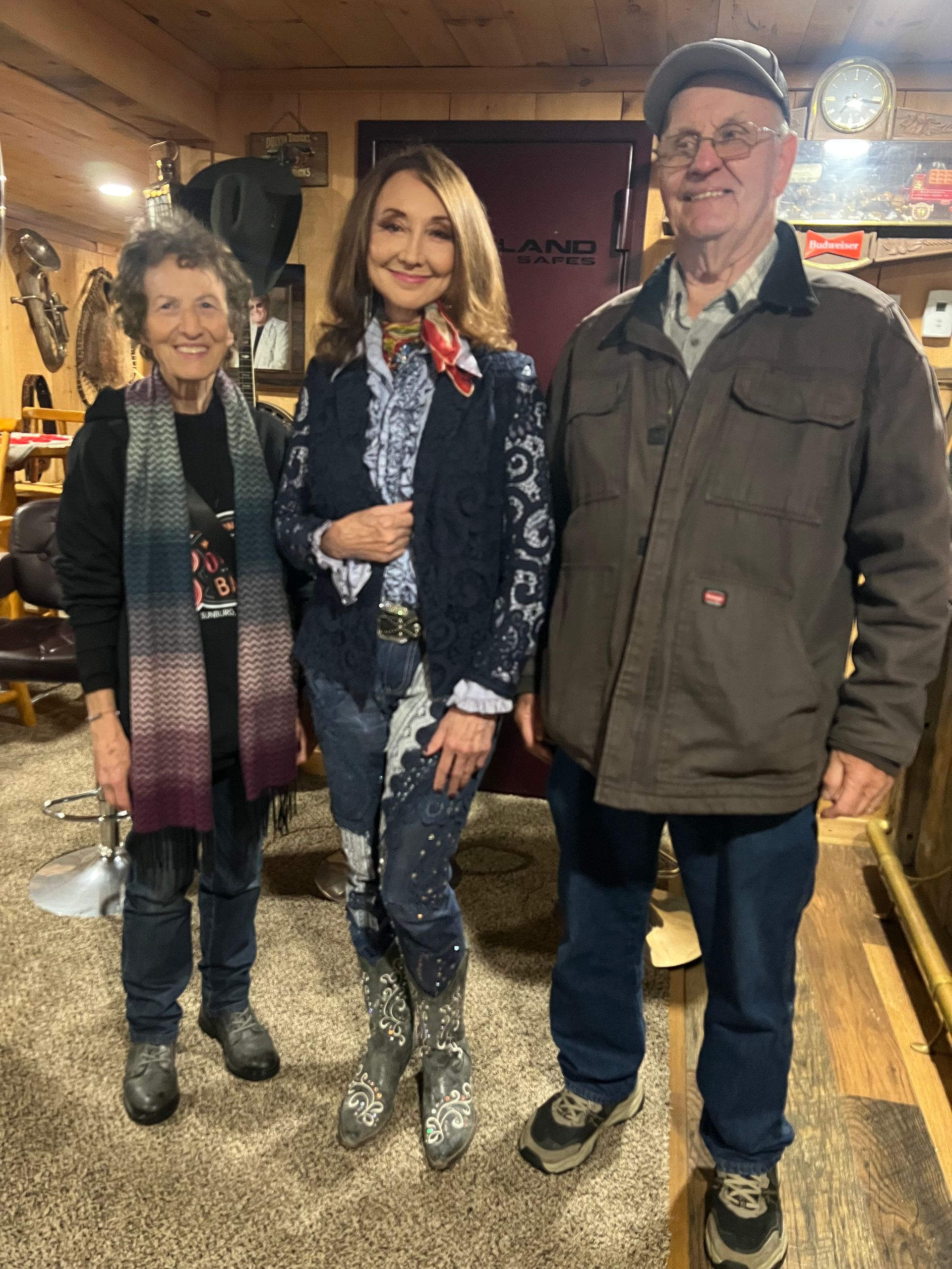 Three people standing indoors, wearing winter coats and boots, smiling at the camera.