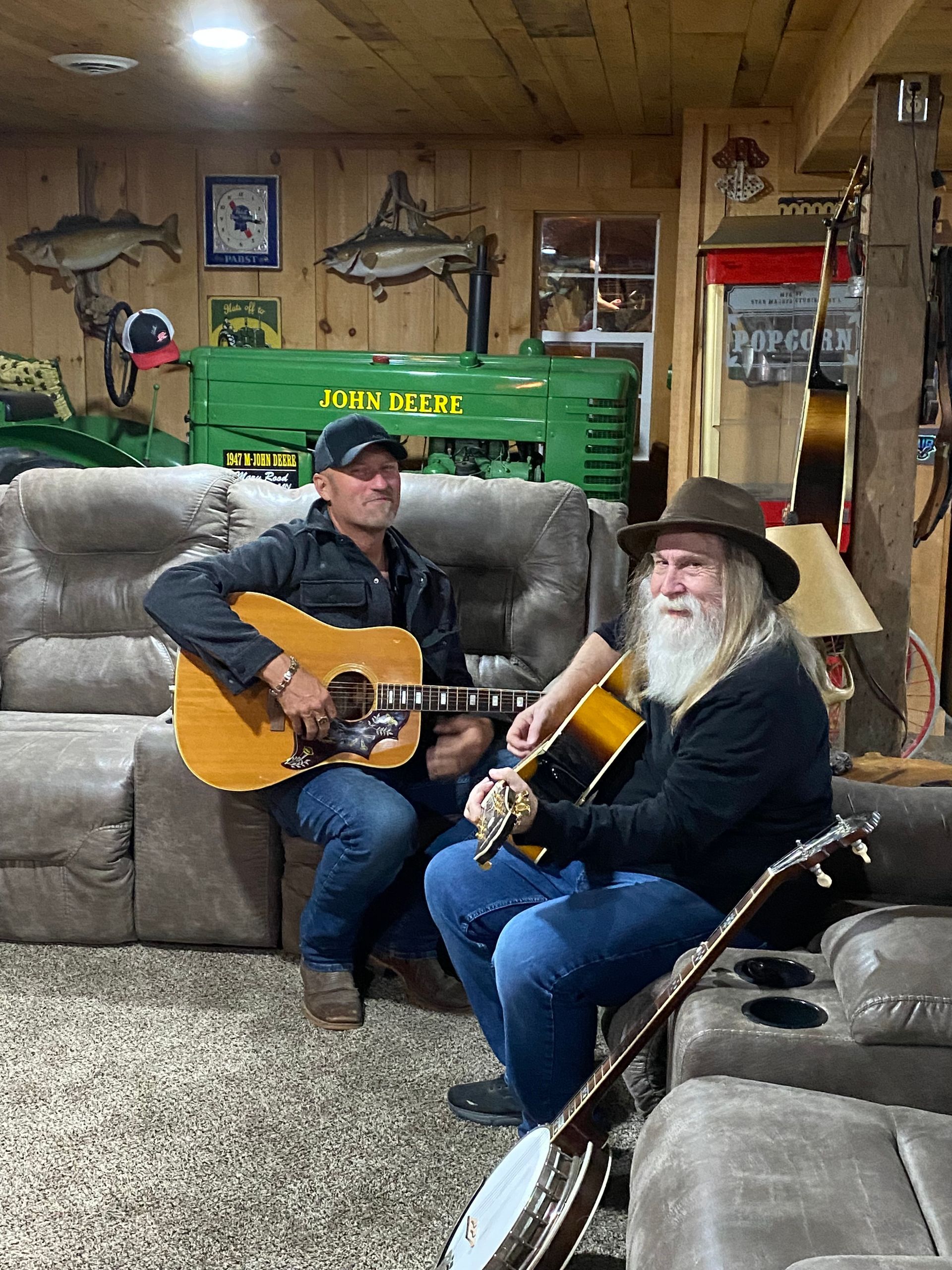 Two people playing guitars in a cozy living room with a couch and wood-paneled walls.