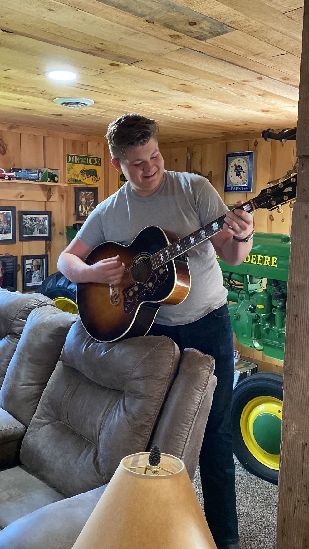 Person playing acoustic guitar in a cozy wood-paneled room with framed photos and a couch in the foreground