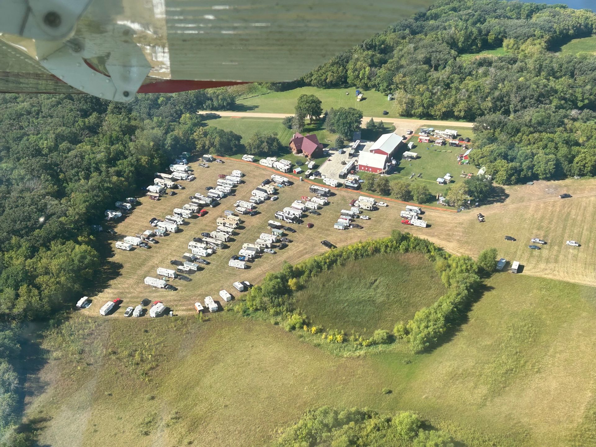 Aerial view of a rural gathering with parked cars, tents, and a pond beside grassy fields and trees.
