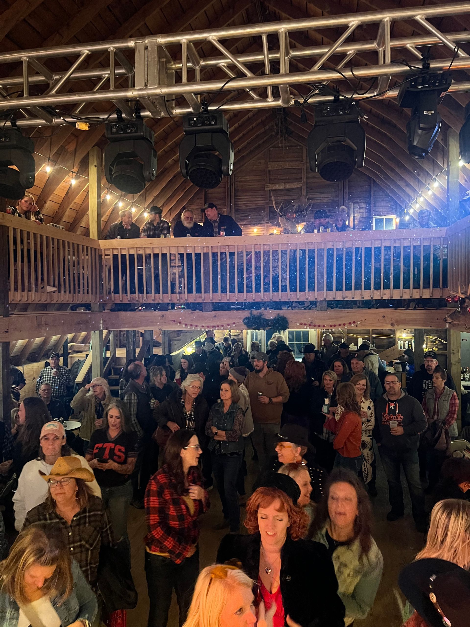 Crowd watching a live performance in a rustic venue with a balcony and stage lights overhead
