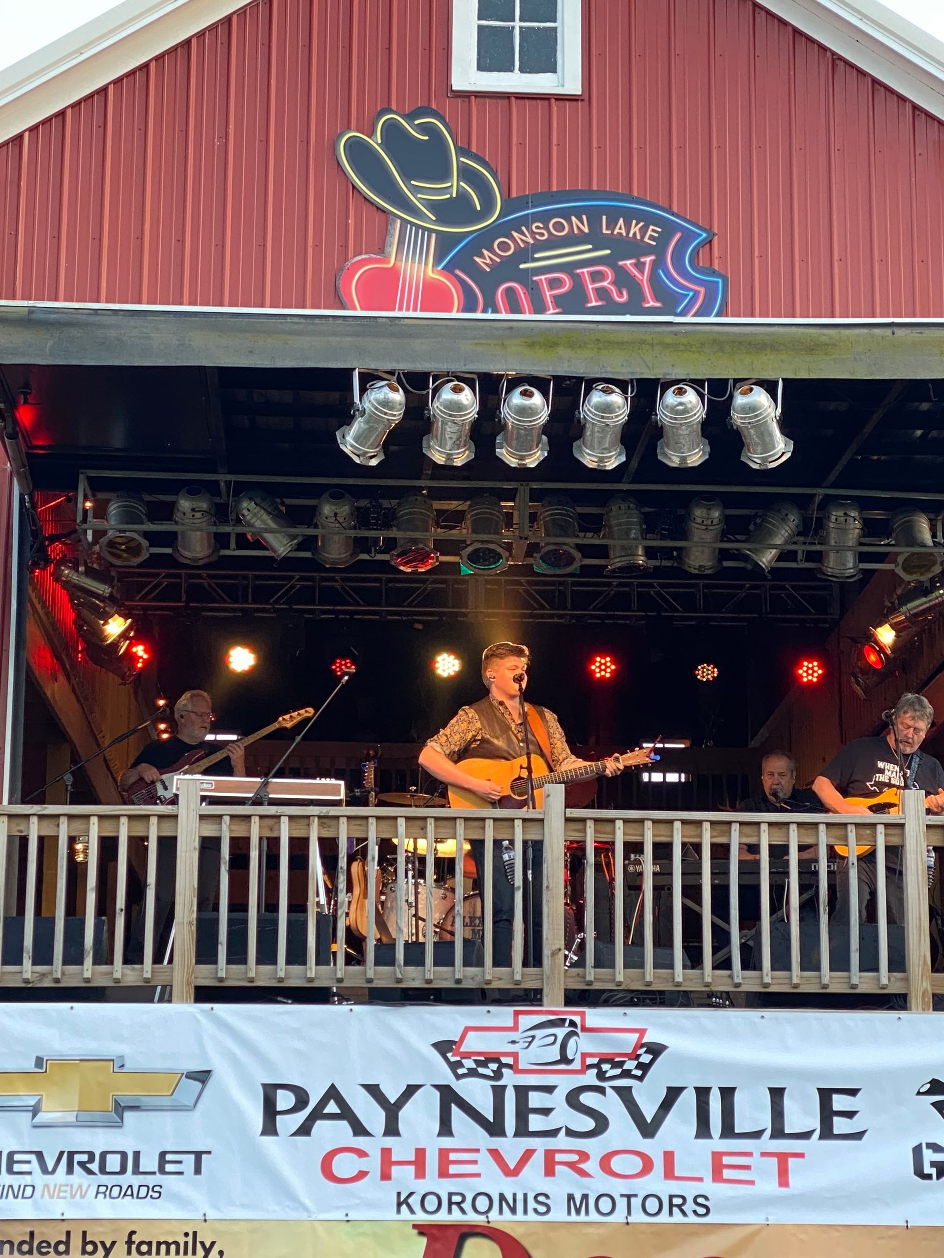 Outdoor stage at a Chevy-sponsored Opry, with musicians performing under lights on a red barn-style backdrop.