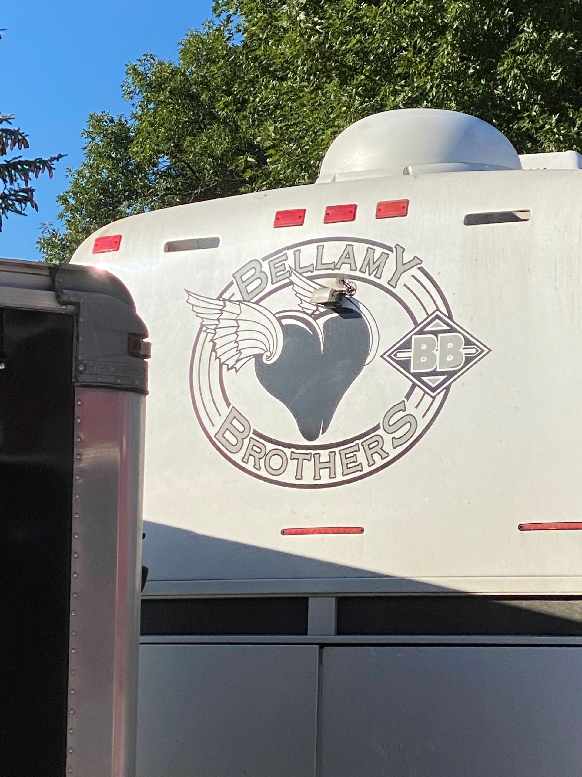 White trailer with a large circular heart logo and red accents, parked outdoors near trees under a blue sky