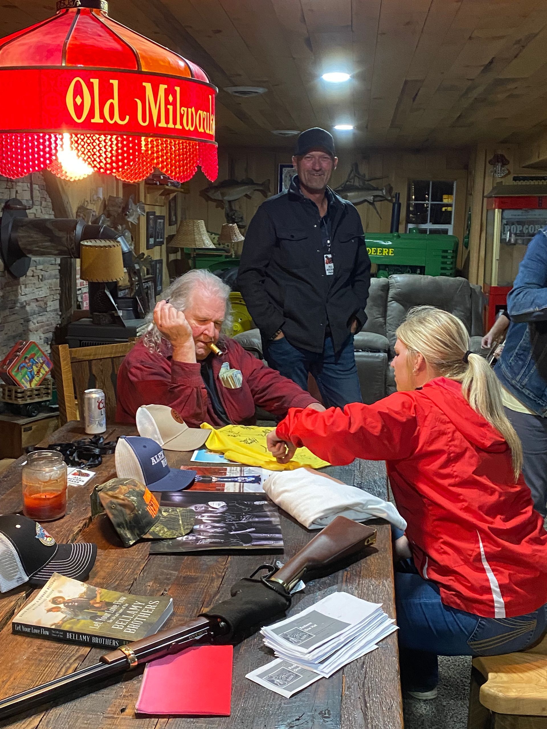 People gathered around a cluttered table under a red lamp in a dim workshop or garage, talking and sorting papers.