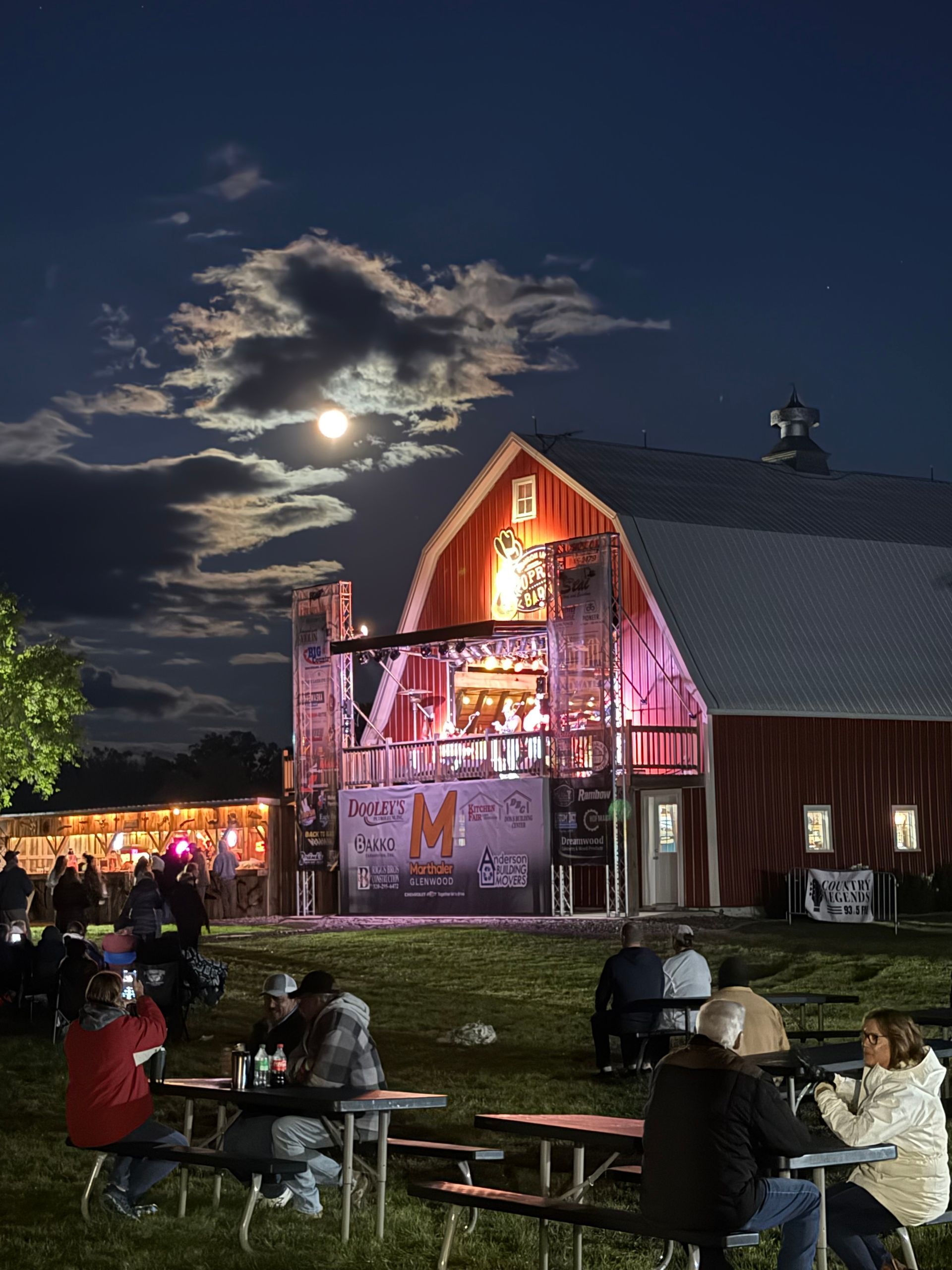 Nighttime outdoor market by a lit barn, with people seated on picnic tables under a moonlit sky.