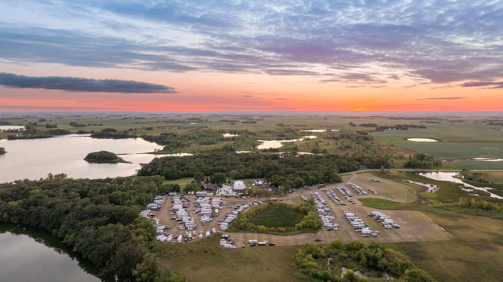 Sunset over a lakeside campground with tents, trees, and a glowing orange-pink sky