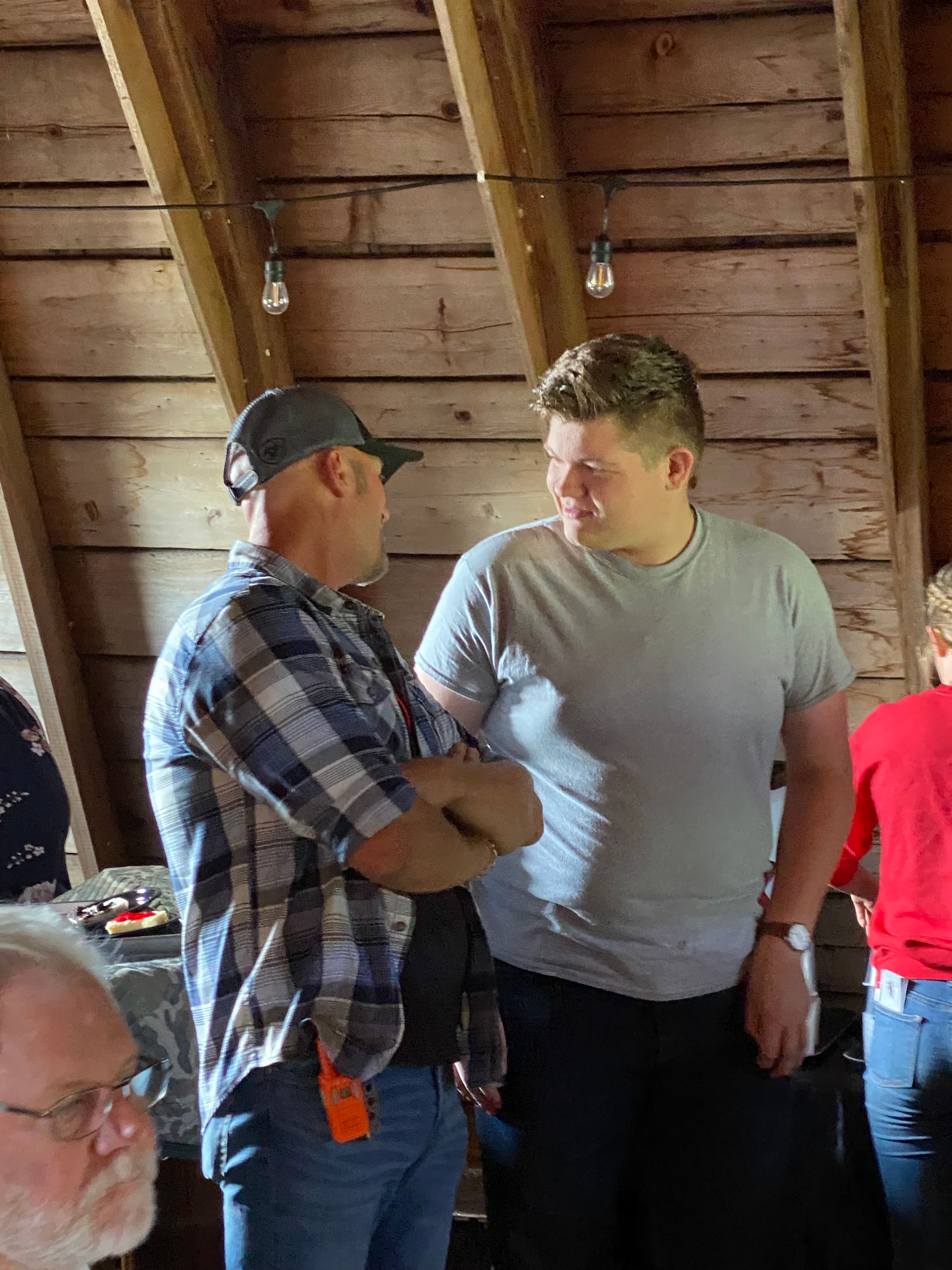 Two men in plaid and gray shirts talking in a wooden barn interior.
