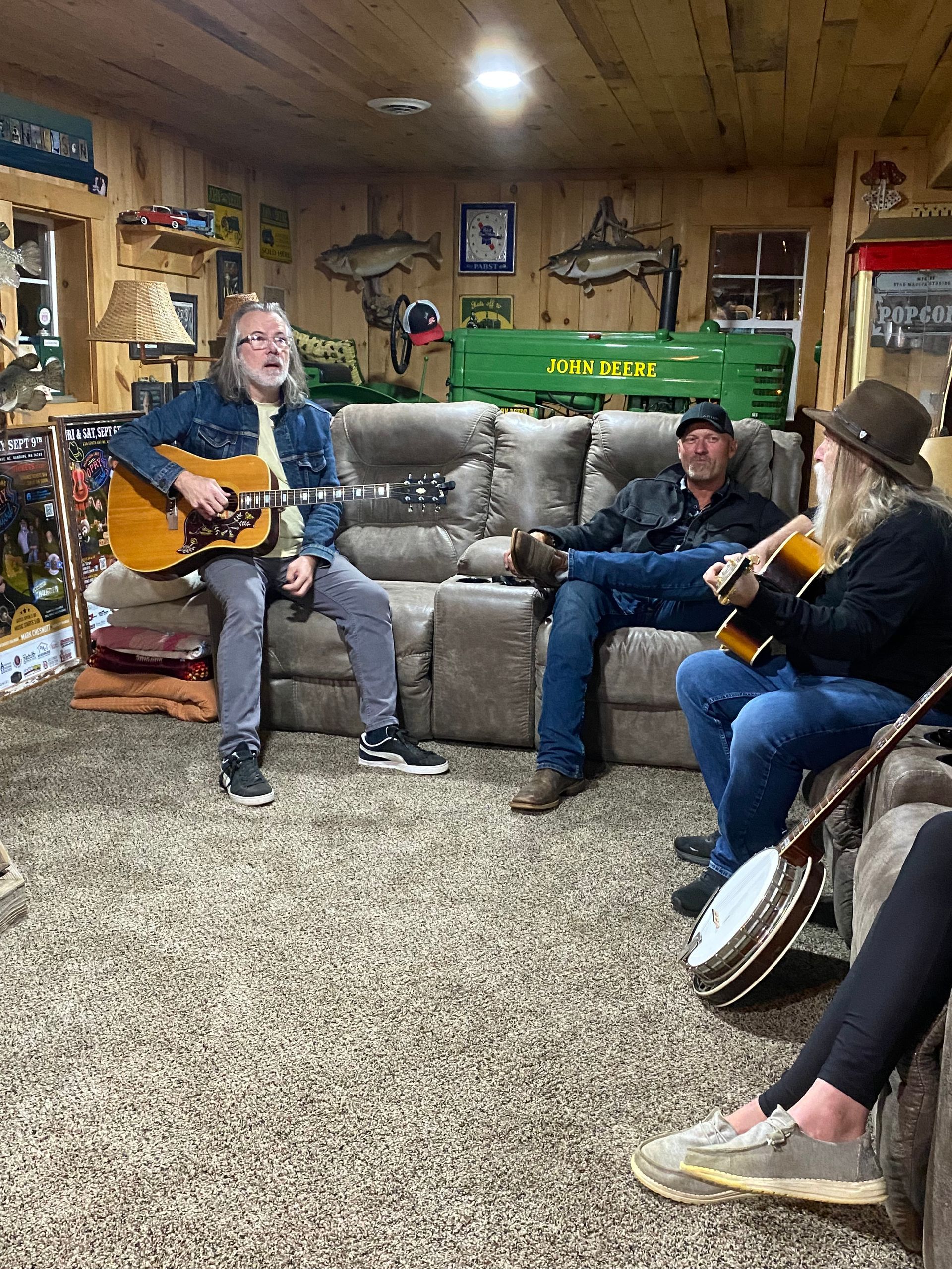 Three people play acoustic guitars in a rustic room with a couch and carpeted floor.