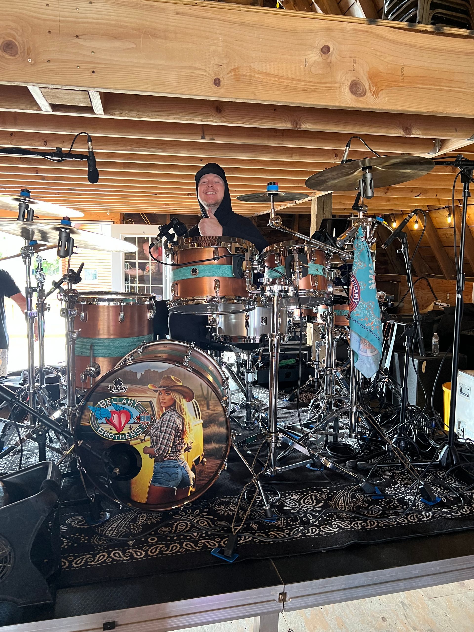 Drummer seated behind a drum kit on a small indoor stage with exposed wooden beams and cymbals overhead