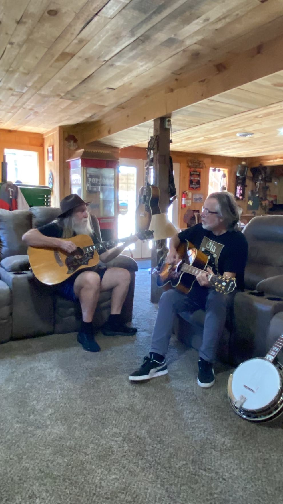 Two people playing guitars on a couch in a cozy cabin living room, with a banjo nearby.