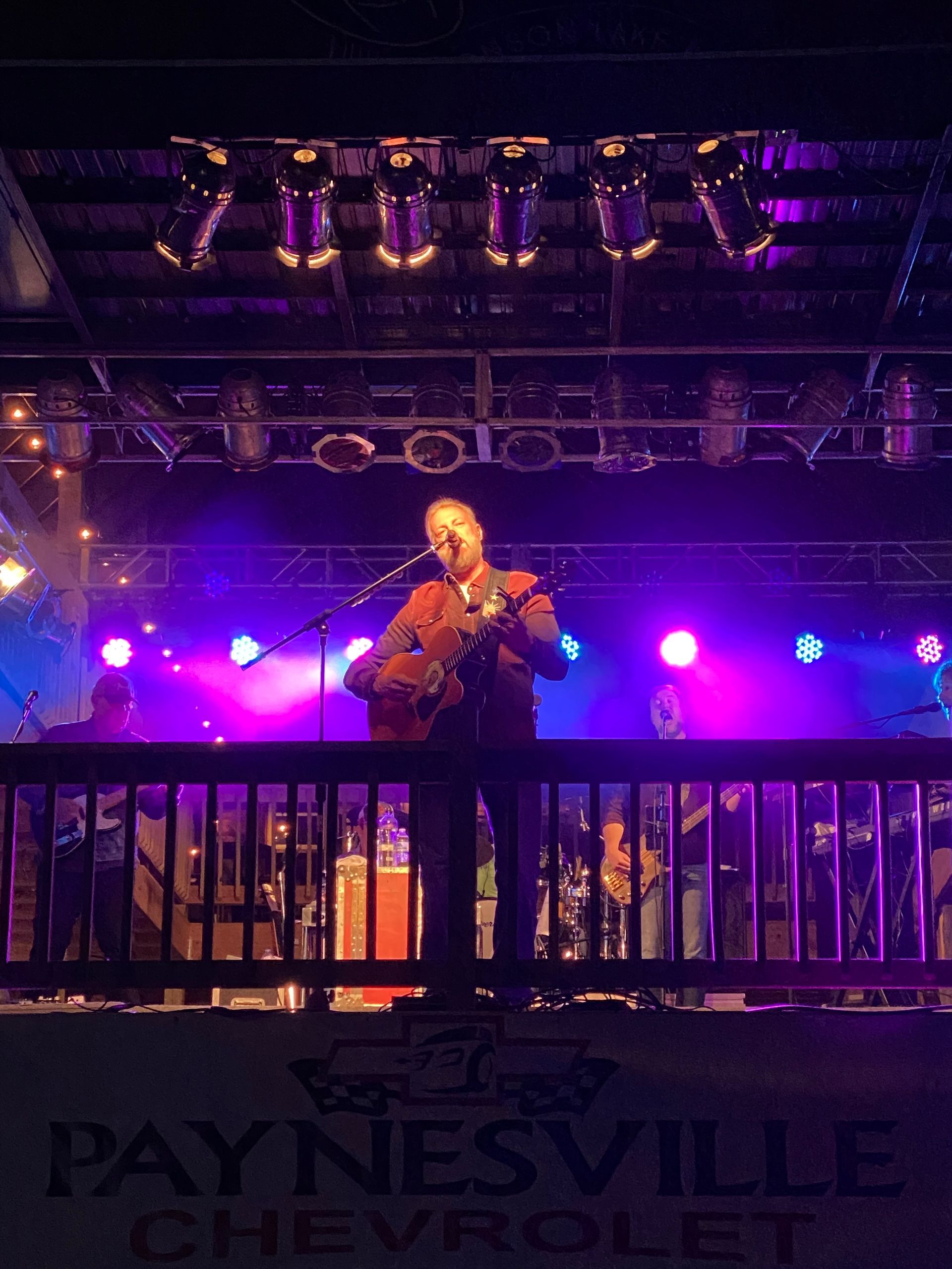 Singer performing on a purple-lit stage behind a railing, with “Paynesville” visible in the foreground.
