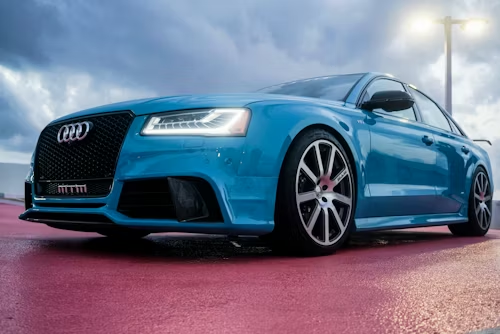 Blue Audi sedan parked on a wet, red surface; dark grill and silver rims; overcast sky.