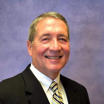 Man in suit smiling at the camera; blue and gold striped tie; purple backdrop.