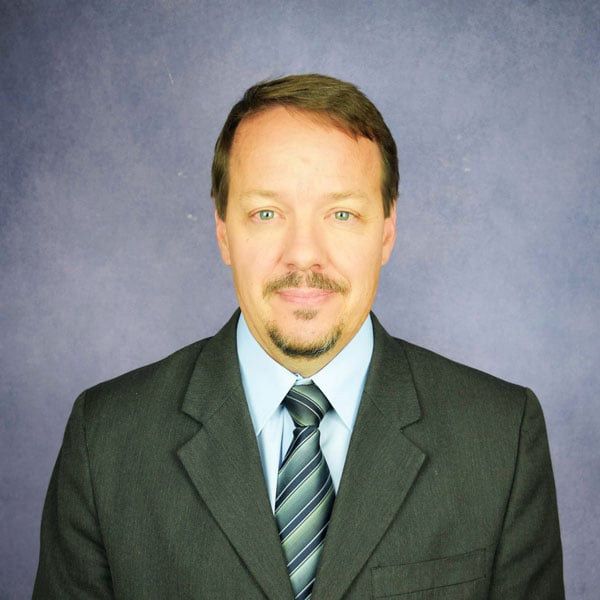 Man in a suit and tie against a blue backdrop; brown hair, mustache, and slight smile.
