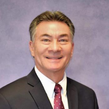 Smiling man in a suit and tie, likely a professional headshot against a purple backdrop.