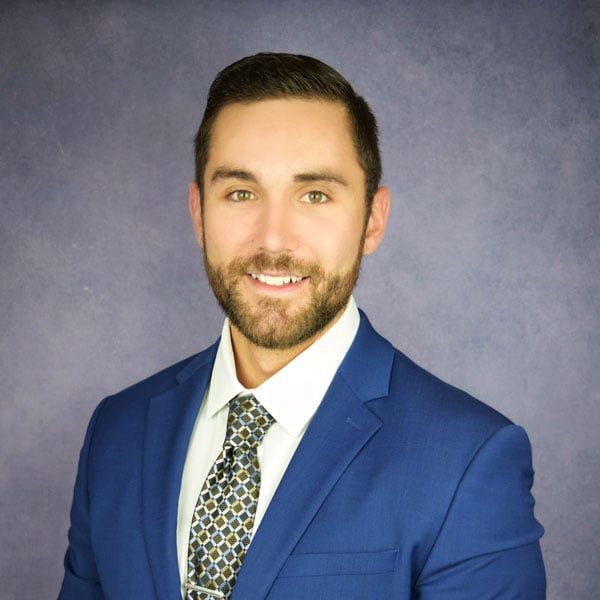 Man in blue suit smiles at the camera, with tie and white shirt against a purple backdrop.