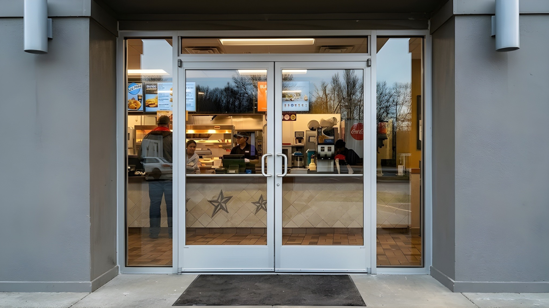 Glass storefront entrance with double doors, indoor lights visible, and a sidewalk mat outside at dusk