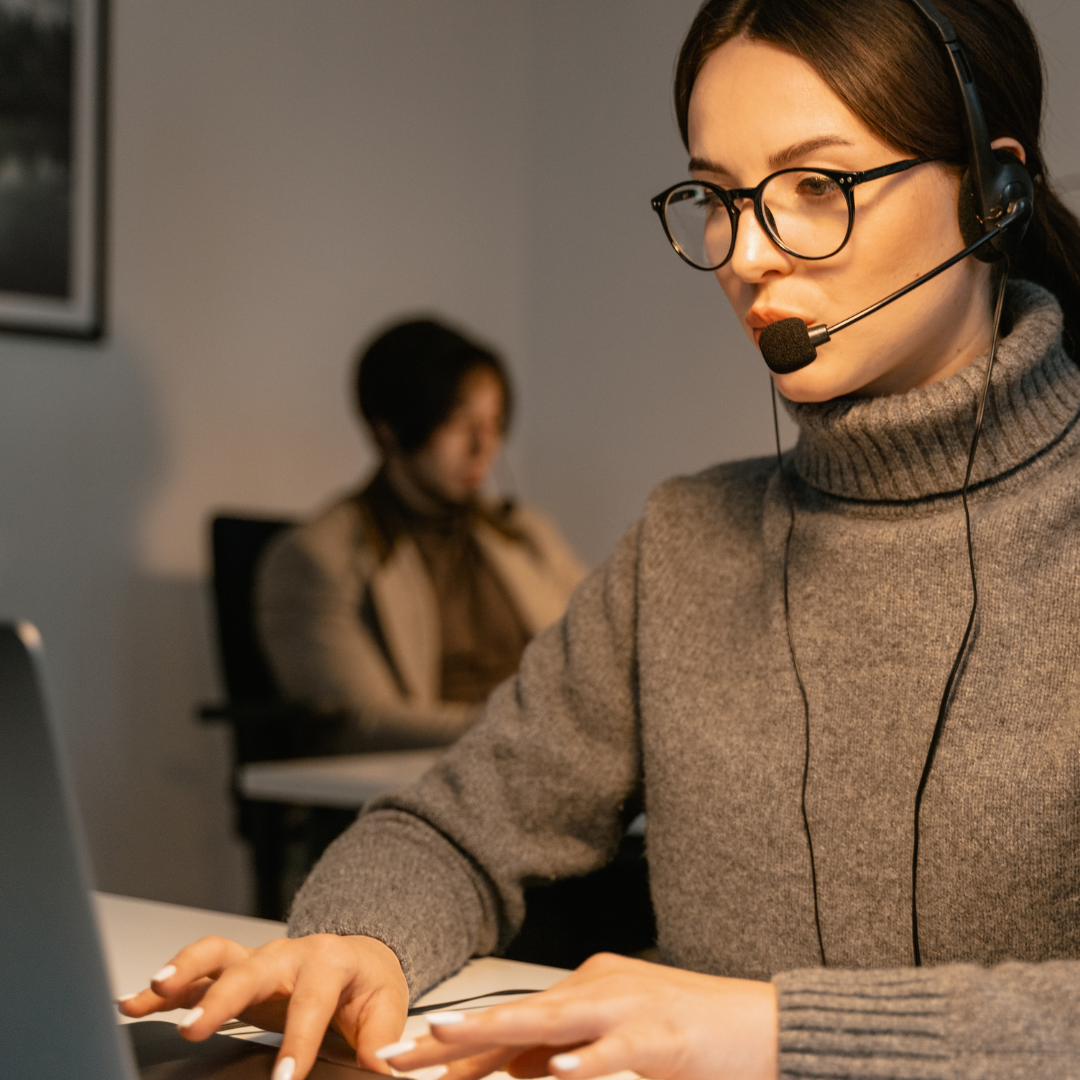 ROK Brothers female employee wearing glasses and headset typing at a laptop in an office, with a blurred coworker in the background