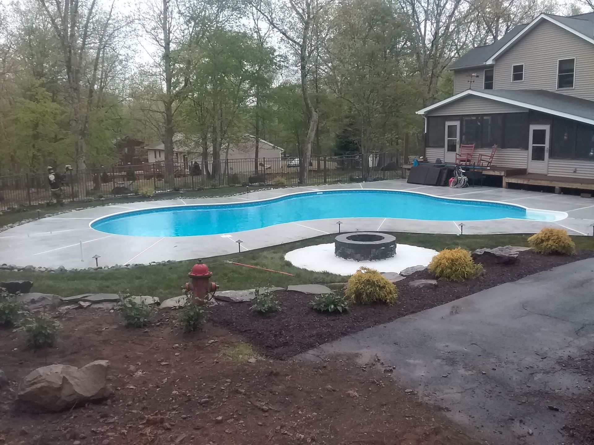 Pool with fire pit in a backyard, house in background, concrete patio, trees and landscaping.