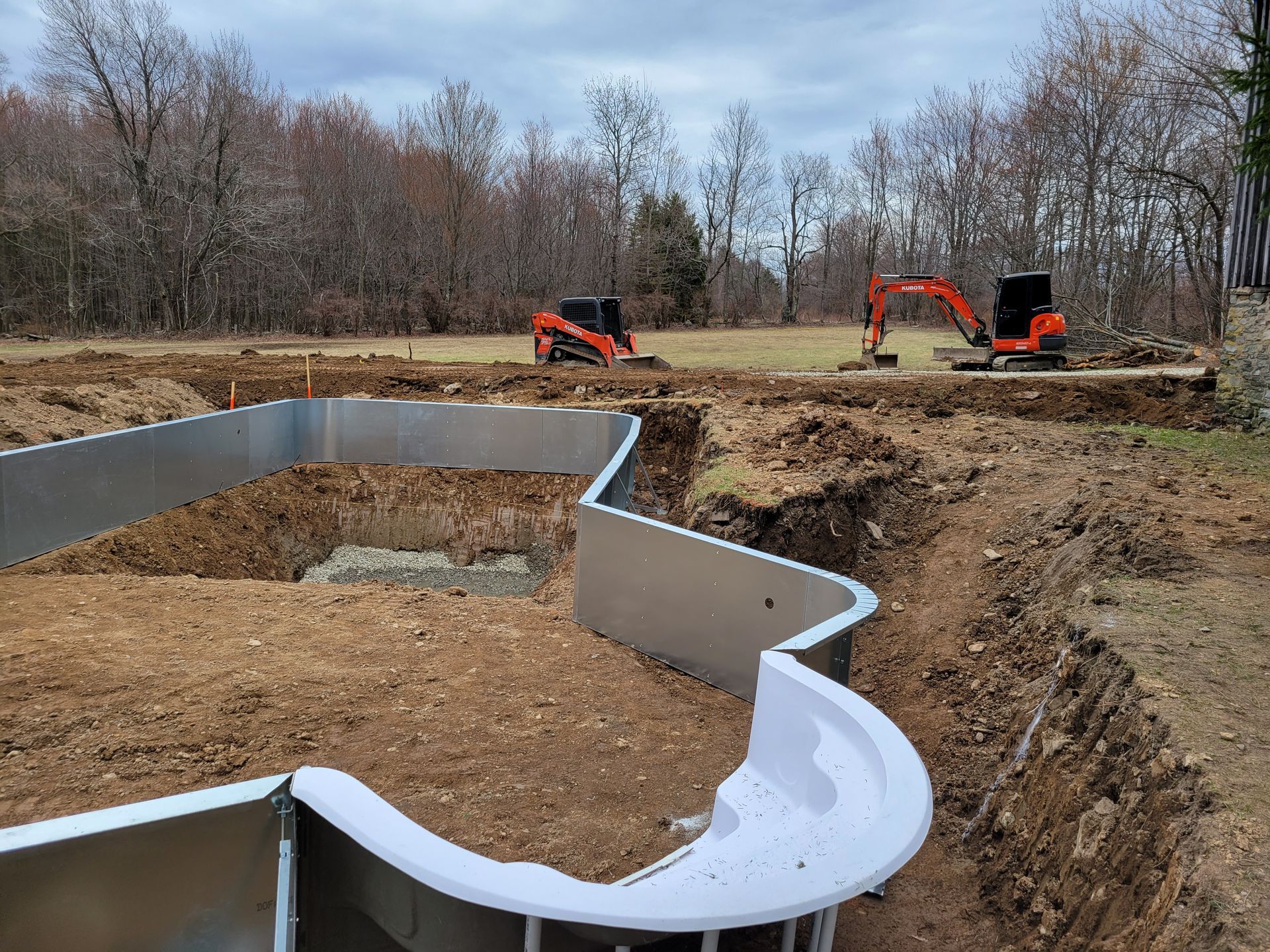 Construction site: Pool excavation with steel walls, machinery, and bare trees.