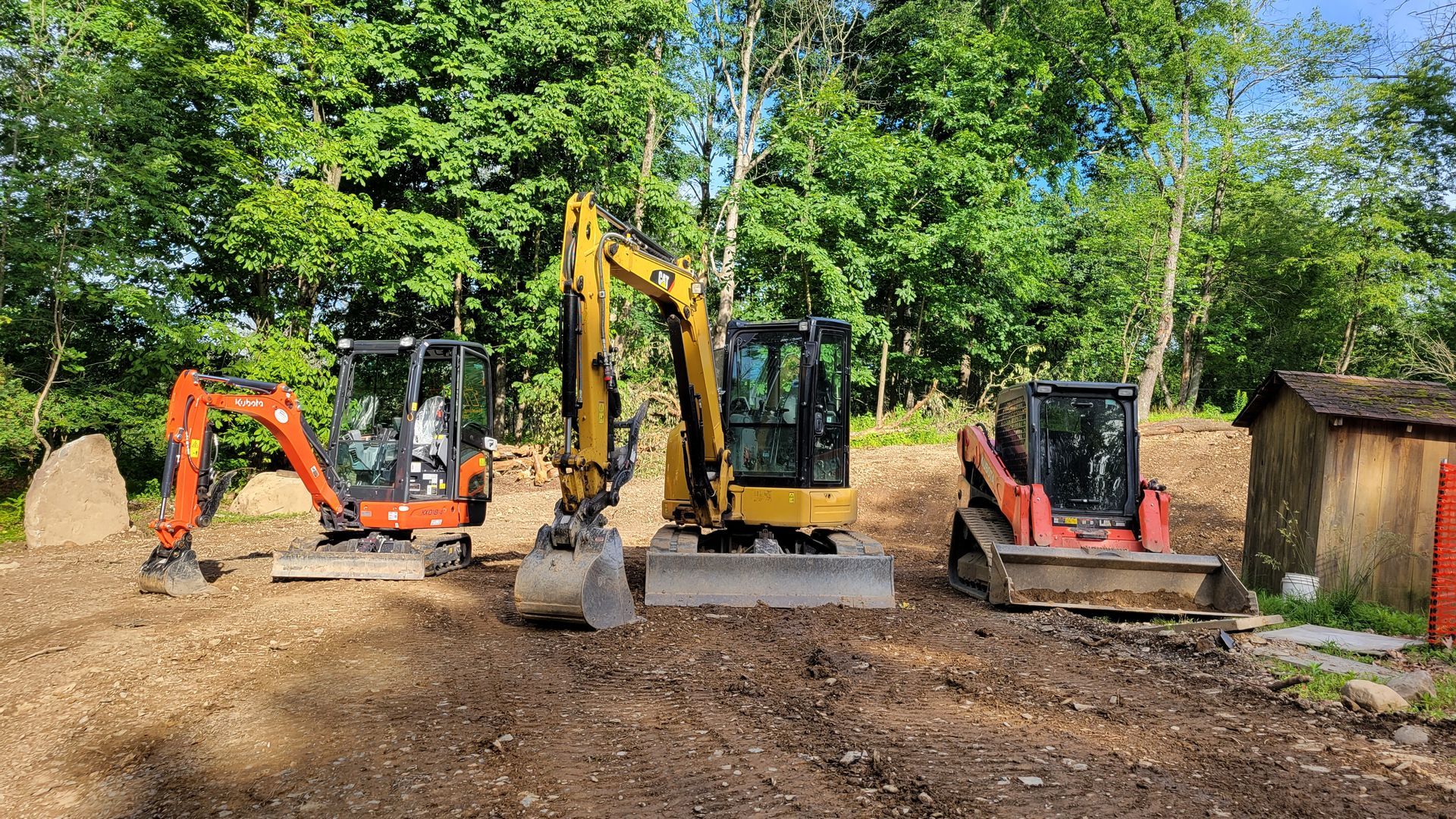 Three construction vehicles: orange excavator, yellow excavator, and red skid steer on a dirt surface.