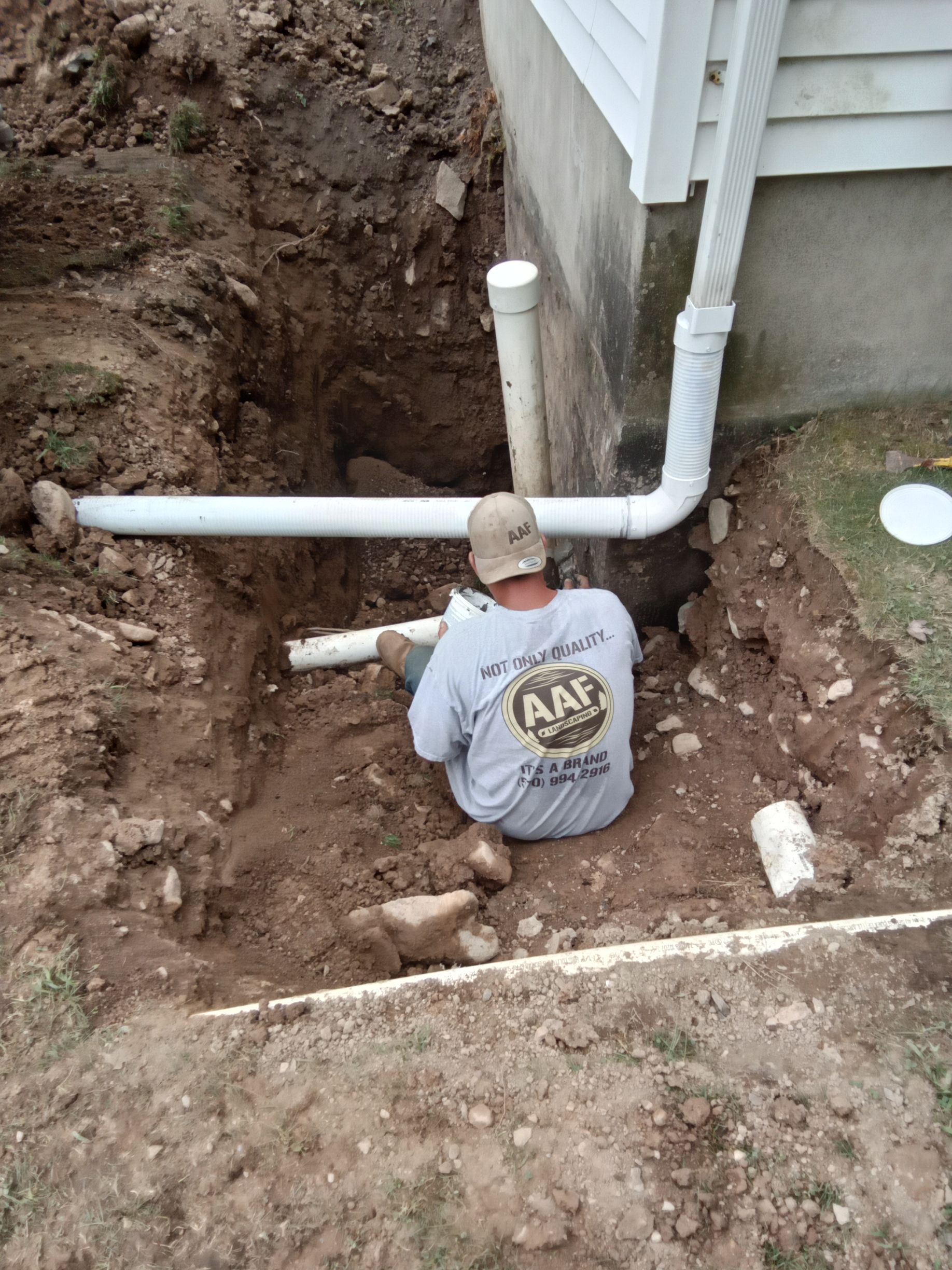 Man installing drainage pipe next to a house foundation. Ground is dug up, white pipe visible.