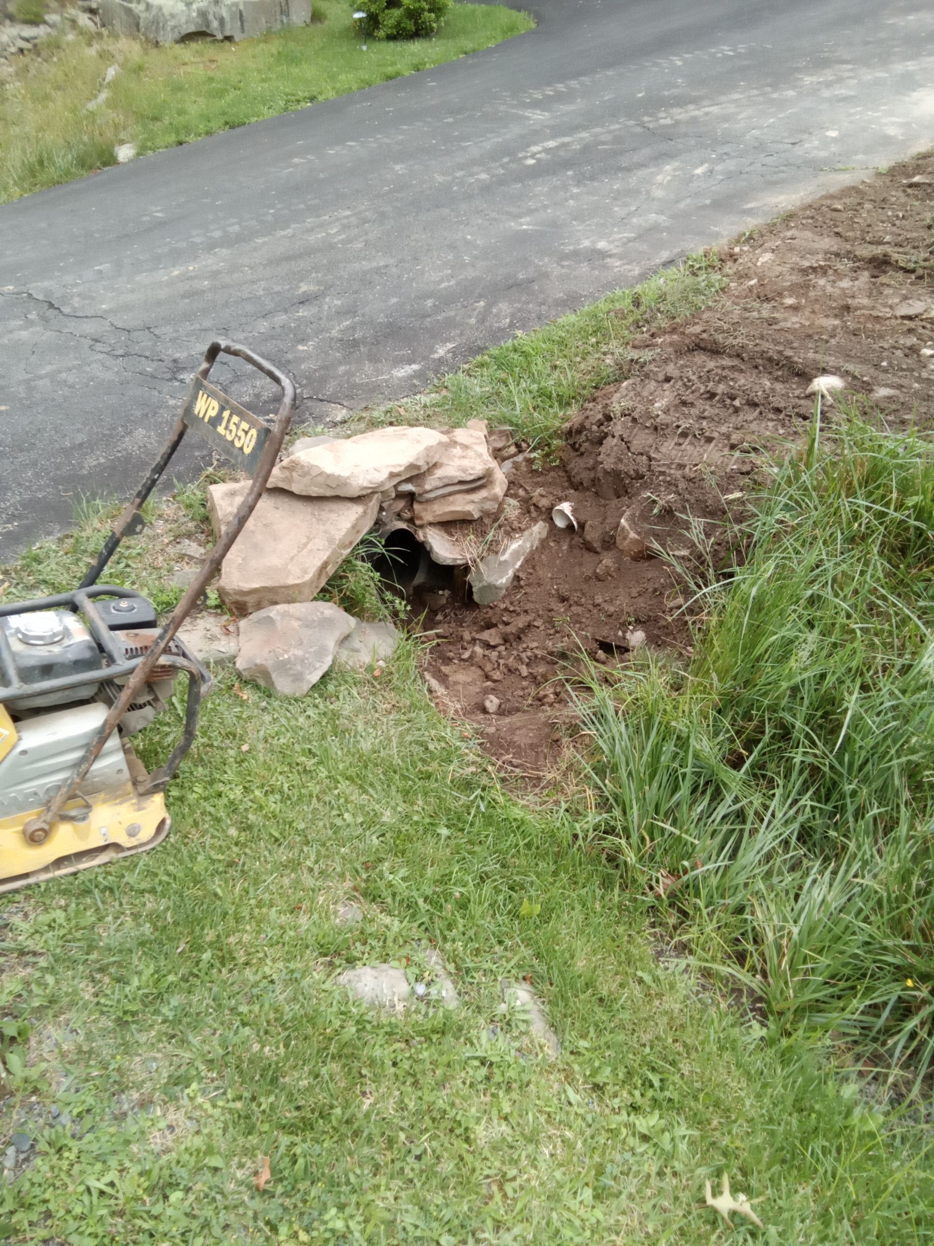 Roadside ditch with soil and rocks, a yellow compactor, and asphalt road.