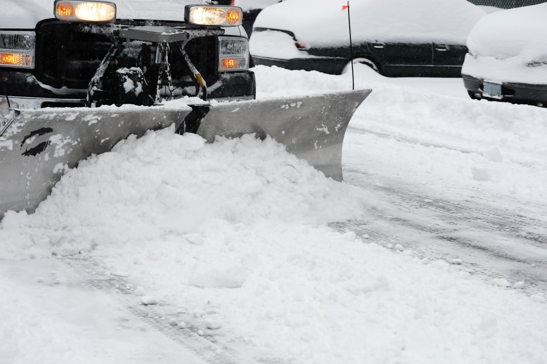 Snowplow clearing snow from a road in a parking lot, gray sky, snow-covered vehicles.