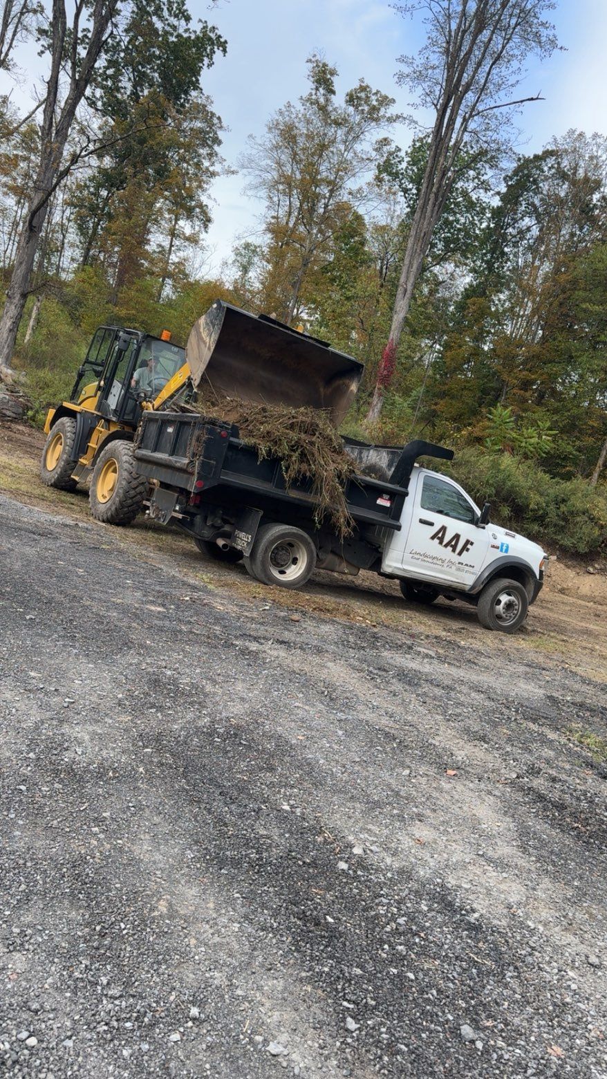 A yellow loader dumping debris into the bed of a white pickup truck. Trees in the background.