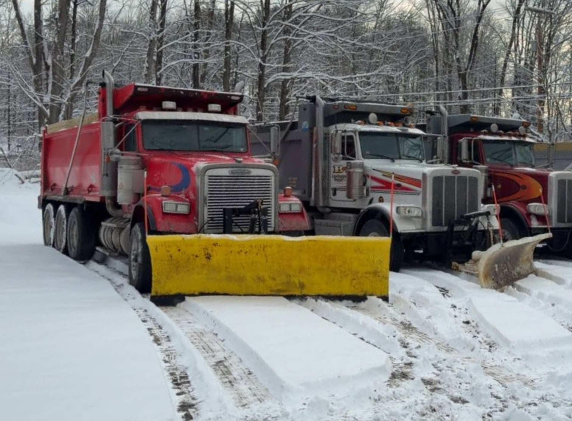 Three snowplow trucks lined up, ready to clear snow. The lead truck is red with a yellow plow.