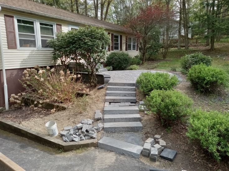 A house with a stone walkway under construction. Steps lead to a porch surrounded by landscaping.