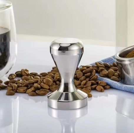Un prensador de café plateado sobre granos esparcidos junto a un vaso y un cuenco sobre una superficie blanca.