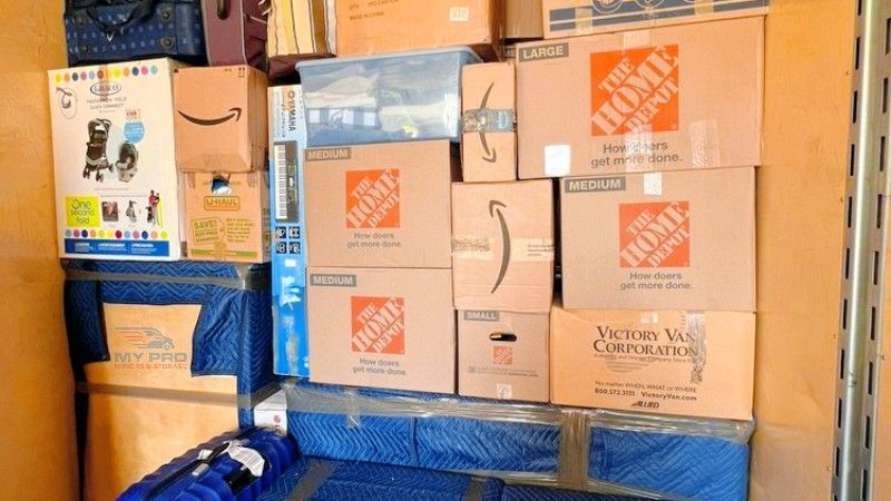 Stacked moving boxes in a storage area, including several Home Depot boxes and blue-wrapped items.