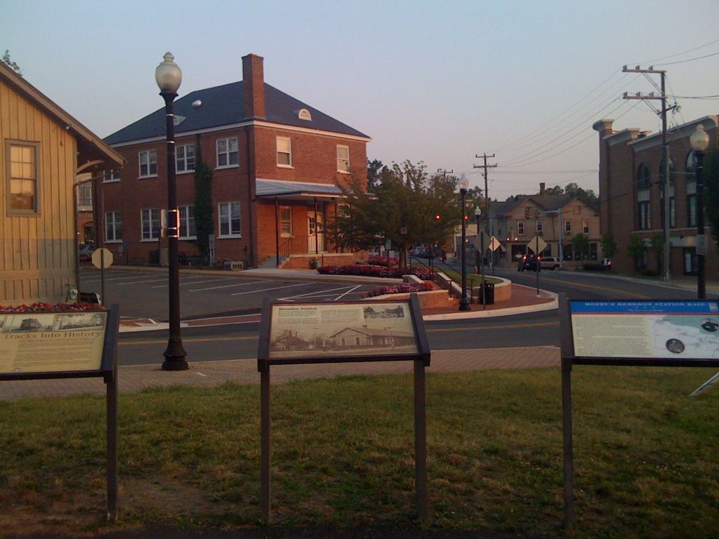 Small-town street with a red brick building, parked cars, lampposts, and signs in the foreground