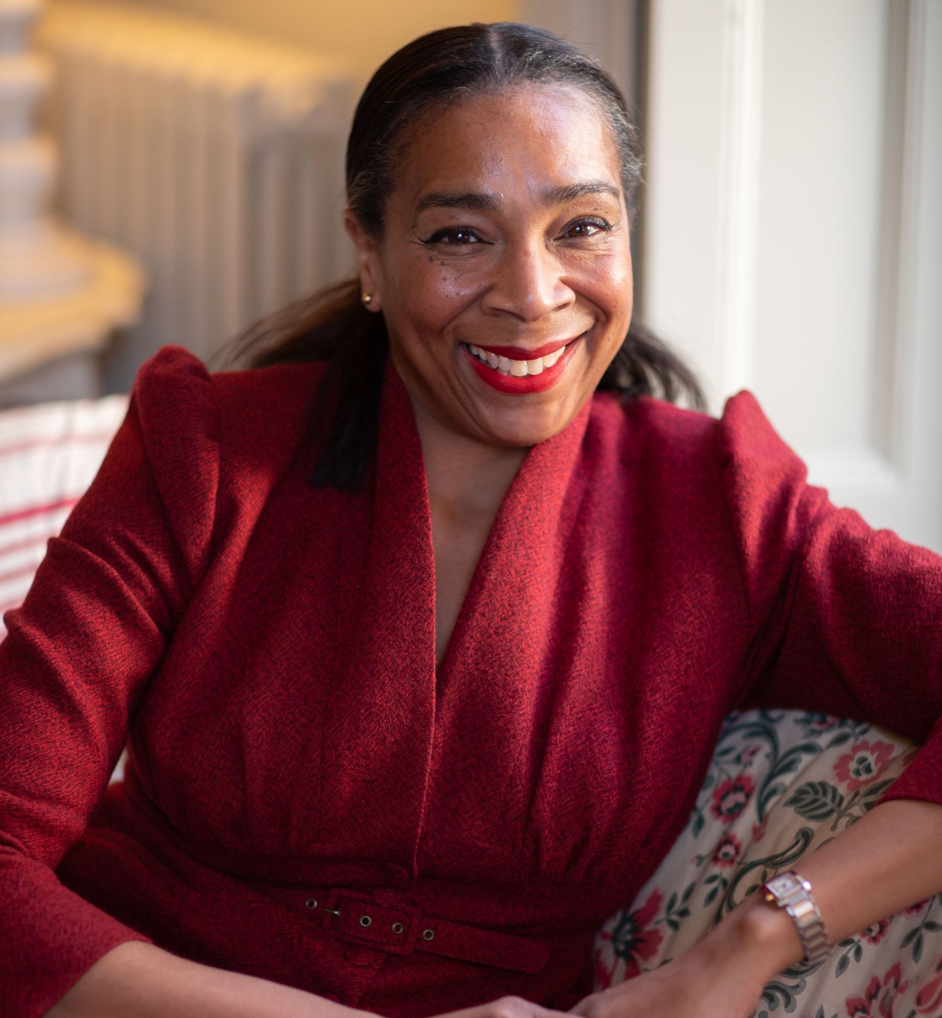 A woman in a red jacket is smiling while sitting on a couch.
