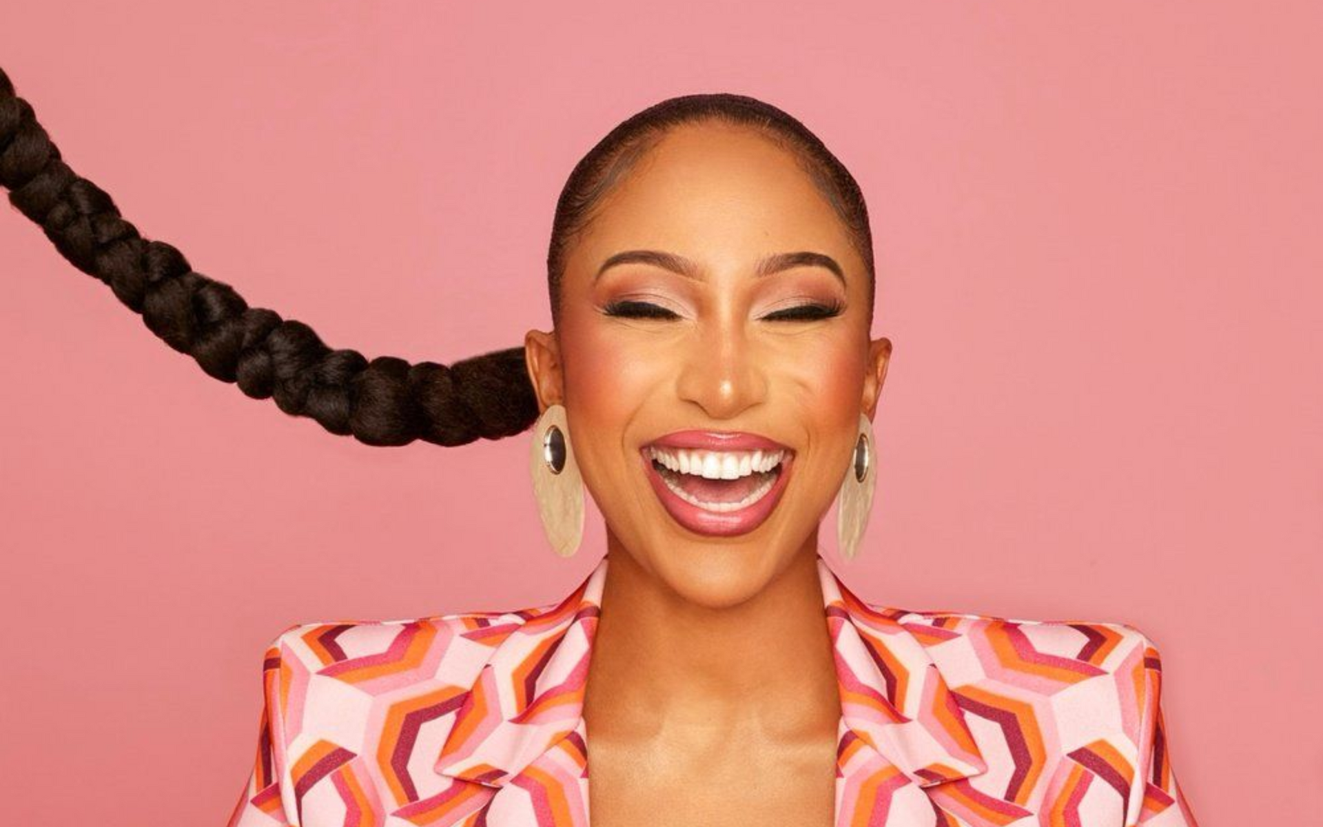 Toni Tone in patterned blazer laughs widely, long braided hair, pink background.