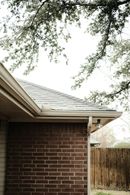 Brick house corner with white eaves, tree branches overhead, and a wooden fence in the background