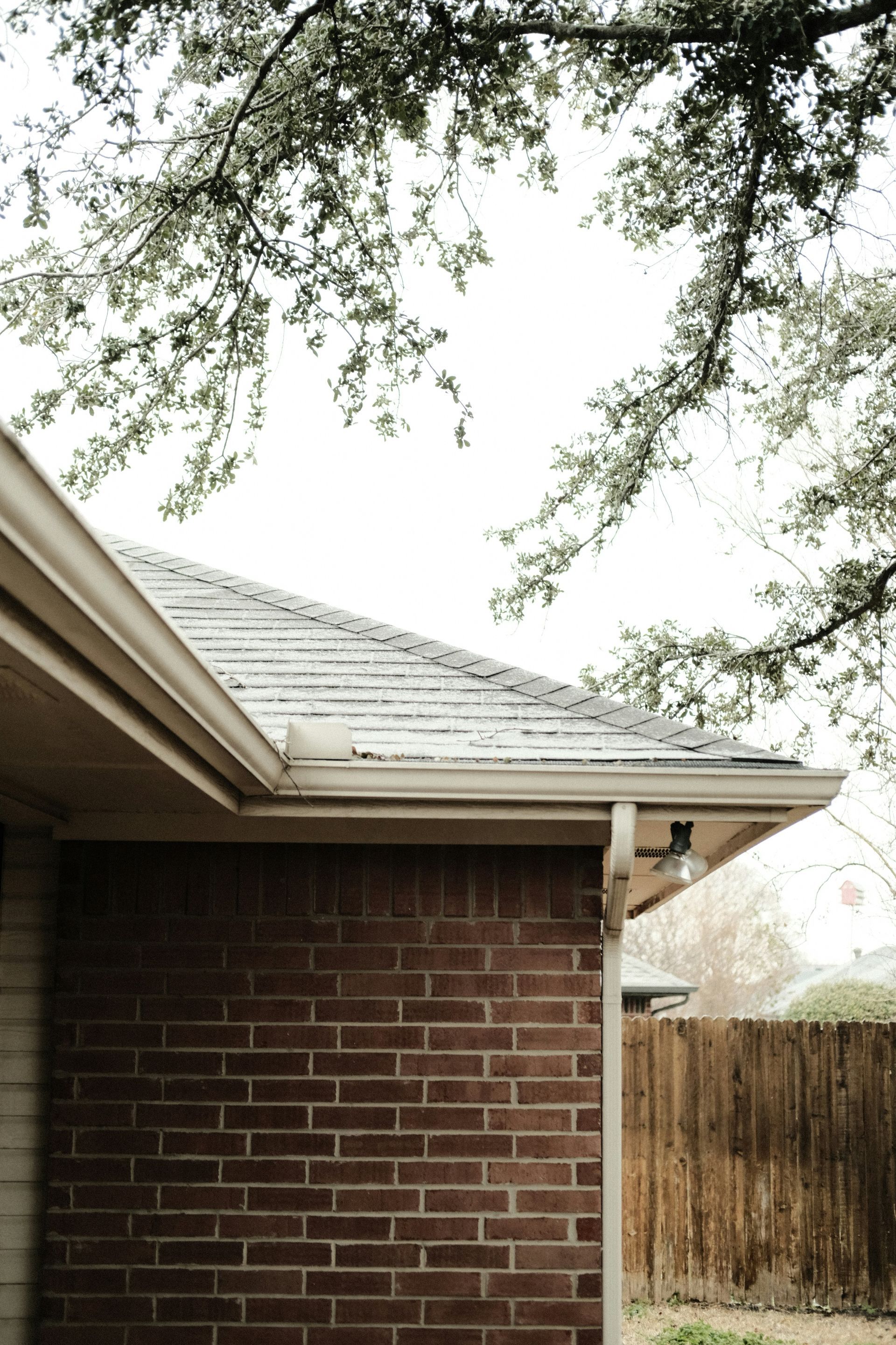 Brick house corner with white eaves, tree branches overhead, and a wooden fence in the background
