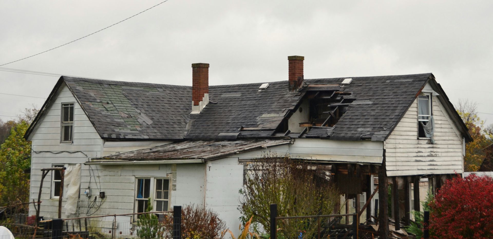 Dilapidated white house with a damaged roof and two chimneys, surrounded by bare shrubs