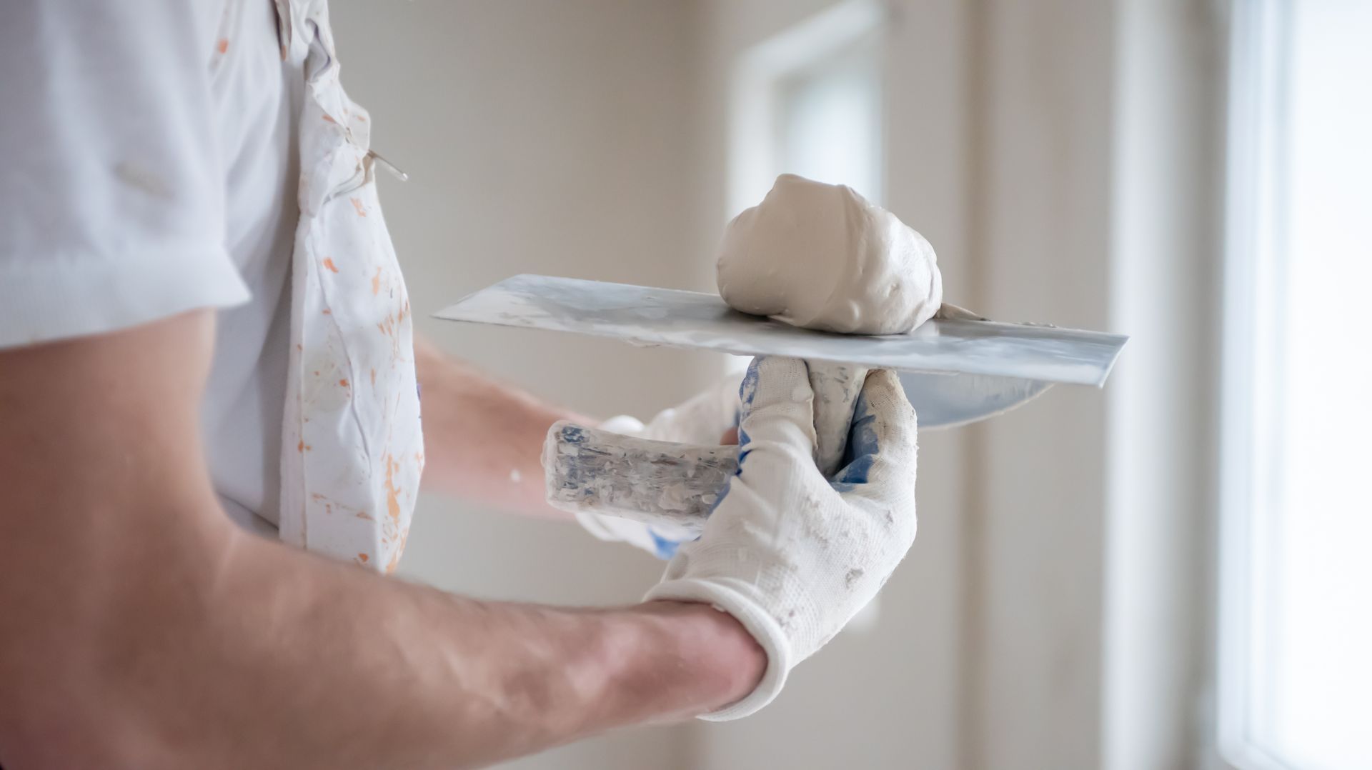 Gloved worker holding a metal trowel with white plaster in a bright room Gloved worker holding a metal trowel with white plaster in a bright room